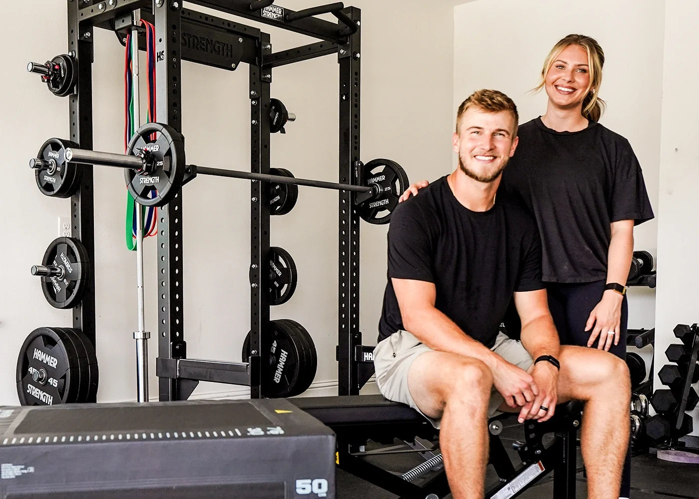 Tyler and Jordan Shough posing in a home gym setting with a black power rack, weight plates, and a bench. The man is sitting on a bench wearing a black t-shirt and khaki shorts, while the woman stands behind him wearing a black t-shirt and dark shorts.