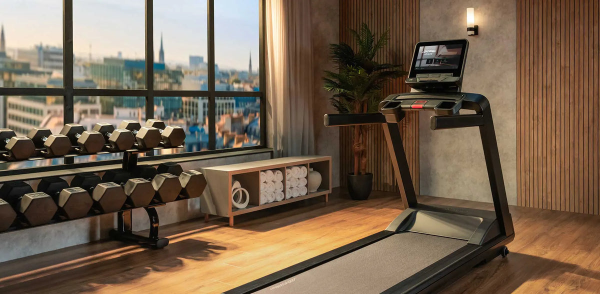 A modern treadmill in a well-lit fitness room with wooden floors, a window view of a city skyline, dumbbells, and a small plant.