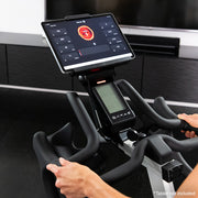 Female holding onto handlebars of ICG Ride CX Indoor Cycle with tablet displaying exercise activity